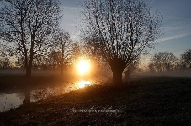 Ein nebliger Sonnenaufgang enthüllt eine heitere Landschaft in Grefrath, wo blattlose Bäume still neben der spiegelnden Oberfläche der Niers im Nebel stehen. HDR-Foto.