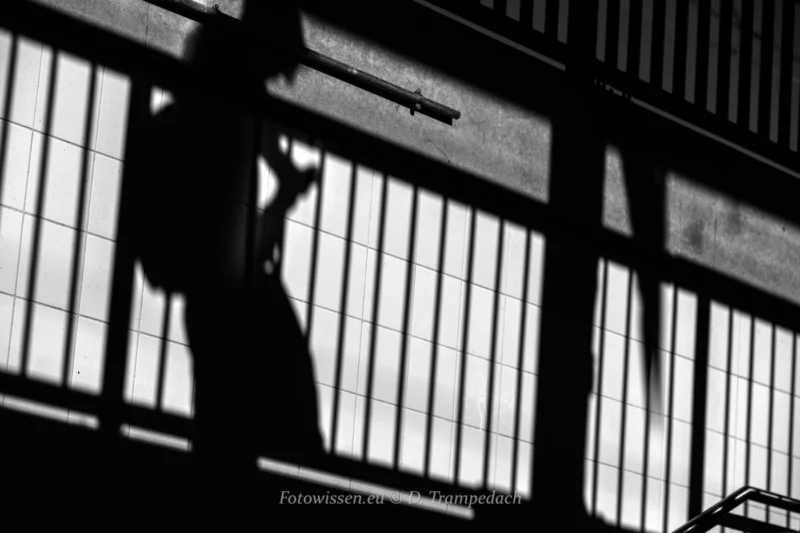 Im Fokus der Street-Fotografie - Das echte Leben -002 Der Schatten einer Person mit Hut und Tasche wandert über eine geflieste Wand und fängt die Essenz des wahren Lebens ein, wie es durch die Linse der Straßenfotografie gesehen wird.